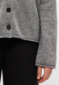 Cardigã cinzento tricotado com um corte solto, apresentando botões escuros, uma borda acabada e textura suave, combinado com calças pretas ajustadas.