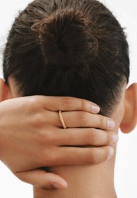 Anillo de oro con un diseño texturizado, llevado en un dedo, mientras una mano reposa suavemente sobre el cuello de una persona, mostrando piel suave y un peinado en moño.