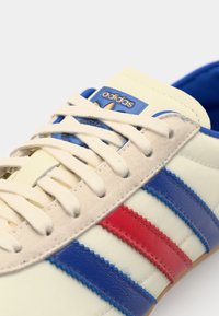 Tenis bej cu dungi albastre și roșii. Prezintă un superior texturat din suede, accente din piele netedă și o etichetă albastră cu un logo aurit.