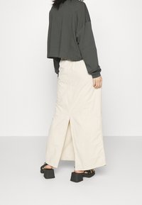 Donna che indossa un top corto scuro a maniche lunghe, una gonna maxi color crema con uno spacco sul retro e sandali platform neri, in piedi contro un muro bianco.