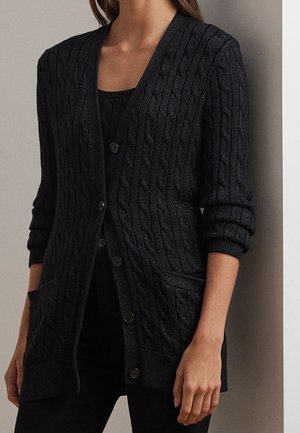 Woman wearing a black cable-knit cardigan with front pockets, button closure, and rolled-up sleeves over a black top.