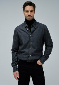 Camisa de mezclilla oscura con cuello abotonado, botones de madera y un sutil logo en el pecho, llevada sobre un cuello de tortuga negro; corte ajustado.