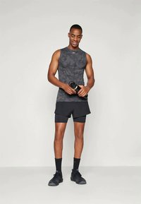 Tumši pelēks bezpiedurknēm sporta krekls ar smalku kamuflāžas rakstu, apvienots ar melniem šortiem un melniem sporta apaviem, tur rokā ūdens pudeli.