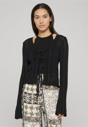 Cardigan nero lavorato a maglia con motivo a rilievo, lunghezza corta, dettaglio con laccio sul davanti e maniche svasate, indossato sopra pantaloni con fantasia.