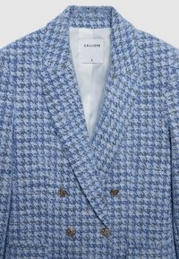 Blazer croisé en tissu pied-de-poule texturé bleu et gris. Présente des boutons métalliques et une étiquette portant l'inscription "CALLIOPE".