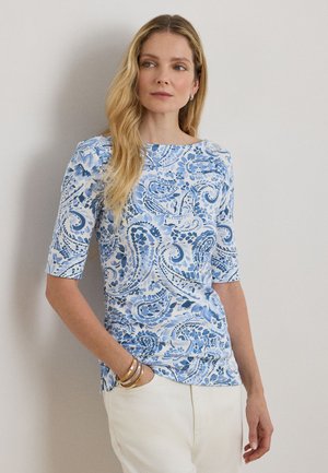 Γυναίκα με μακριά ξανθά μαλλιά φορώντας πάνω μέρος με μπλε και λευκά σχέδια παϊέτας και λευκό παντελόνι, όρθια με το ένα χέρι στην τσέπη.