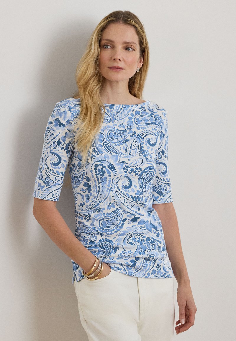 Femme aux longs cheveux blonds portant un haut à motif cachemire bleu et blanc et un pantalon blanc, debout avec une main dans la poche.