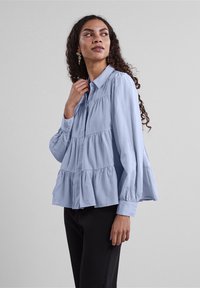 Himmelblaue Bluse mit langen Ärmeln, Knopfverschluss, gestuftem Rüschendesign und Kragen. Glattes Material und lockere Passform.