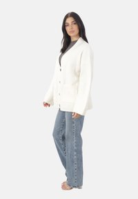 Kobieta z długimi ciemnymi włosami, ubrana w białą, oversize'ową kardiganę, szary t-shirt, jasnoniebieskie dżinsy i przezroczyste obcasy, stojąca na białym tle.