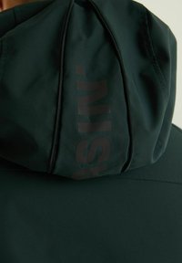 Zelena kapuca z jopico iz mehkega, teksturiranega blaga; ima zadrgo in velik mat črn logo na kapuci.