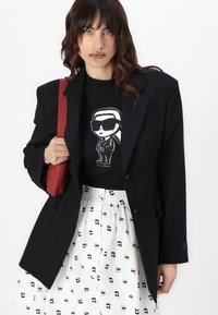 Svart overdimensjonert blazer med strukturerte skuldre, brukt over en grafisk svart t-skjorte med en tegneseriefigur, kombinert med et hvitt skjørt med svarte mønstre.