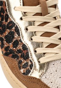Primer plano de una zapatilla con cordones beige, ante marrón, ribete metálico dorado y un panel de estampado de leopardo con lentejuelas negras y marrones.