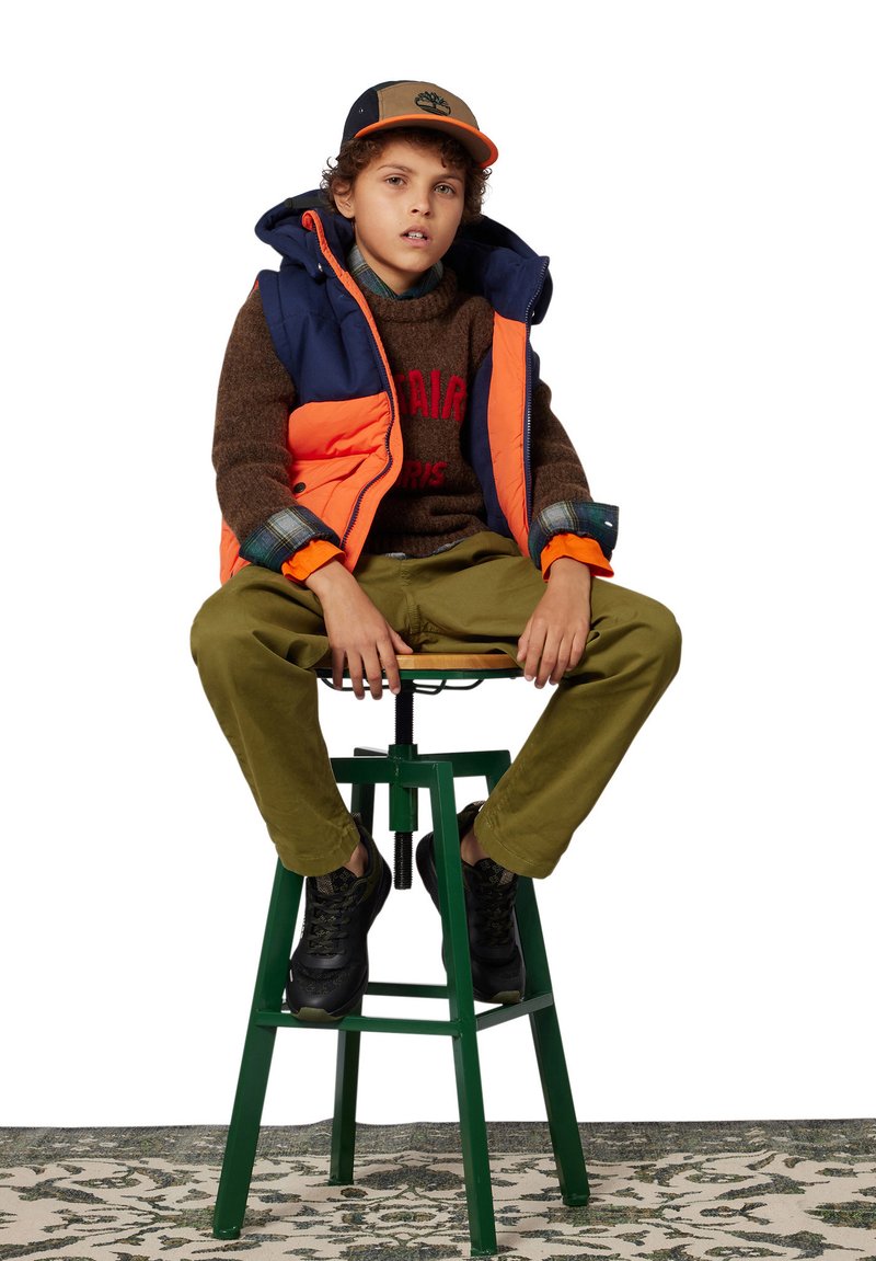 Gilets rembourrés en bleu marine et orange, pull brun avec texte rouge, pantalons verts et baskets noires. Enfant assis sur un tabouret vert.