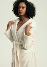 Miękki kremowy szlafrok z polaru, wyposażony w pasek, głębokie kieszenie i kołnierz szalowy; gładka faktura i luźny krój.