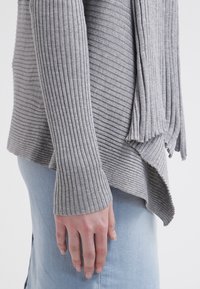 Main reposant le long du corps portant un pull à manches longues gris côtelé et une jupe en denim bleu clair avec un ourlet asymétrique.