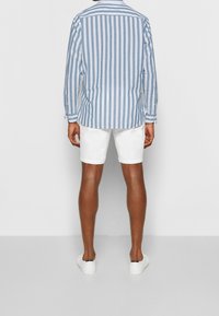Camicia button-up a righe blu chiaro e bianche, maniche lunghe, indossata con pantaloni corti bianchi e sneaker bianchi, vista da dietro.