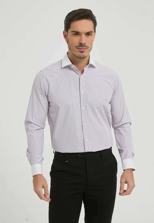 Homme portant une chemise blanche à fines rayures verticales et poignets blancs, associée à un pantalon noir, debout devant un fond uni.