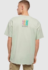 Lys grøn oversized t-shirt med korte ærmer, med en flerfarvet tekstgrafik "PRIDE SCOOPS" på ryggen. Klassisk rund halsudskæring.