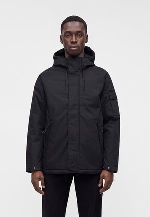 Homme portant une veste noire à capuche avec des poches zippées et des poignets boutonnés, se tenant devant un fond clair et uni.