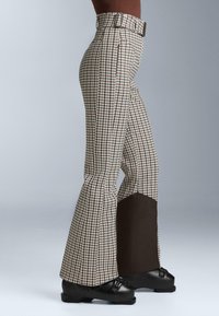 Vysoké páskované nohavice so vzorom houndstooth, ktoré sa na spodnej časti nohavíc rozširujú, so sivými panelmi na dolnej časti nôh, nosené spolu s čiernymi snehovými čižmami, na osobe proti šedému pozadiu.