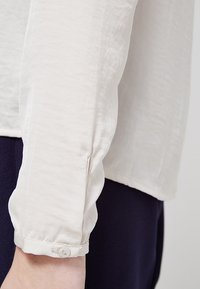 Camicia beige chiaro a maniche lunghe con un tessuto testurizzato e dettaglio dei polsini con bottoni, contrastata su uno sfondo scuro.