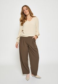 Žena s dlhými blond vlasmi, oblečená v béžovom topánke s dlhým rukávom, hnedých širokých nohaviciach, béžových lodičkách a hnedom náramku, stojaca s jednou rukou v vrecku.