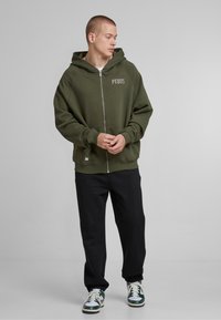 Sudadera con cremallera de color verde oliva, con una textura suave, mangas raglán y un logo en la parte frontal. Combinada con pantalones negros y zapatillas con acentos en verde.