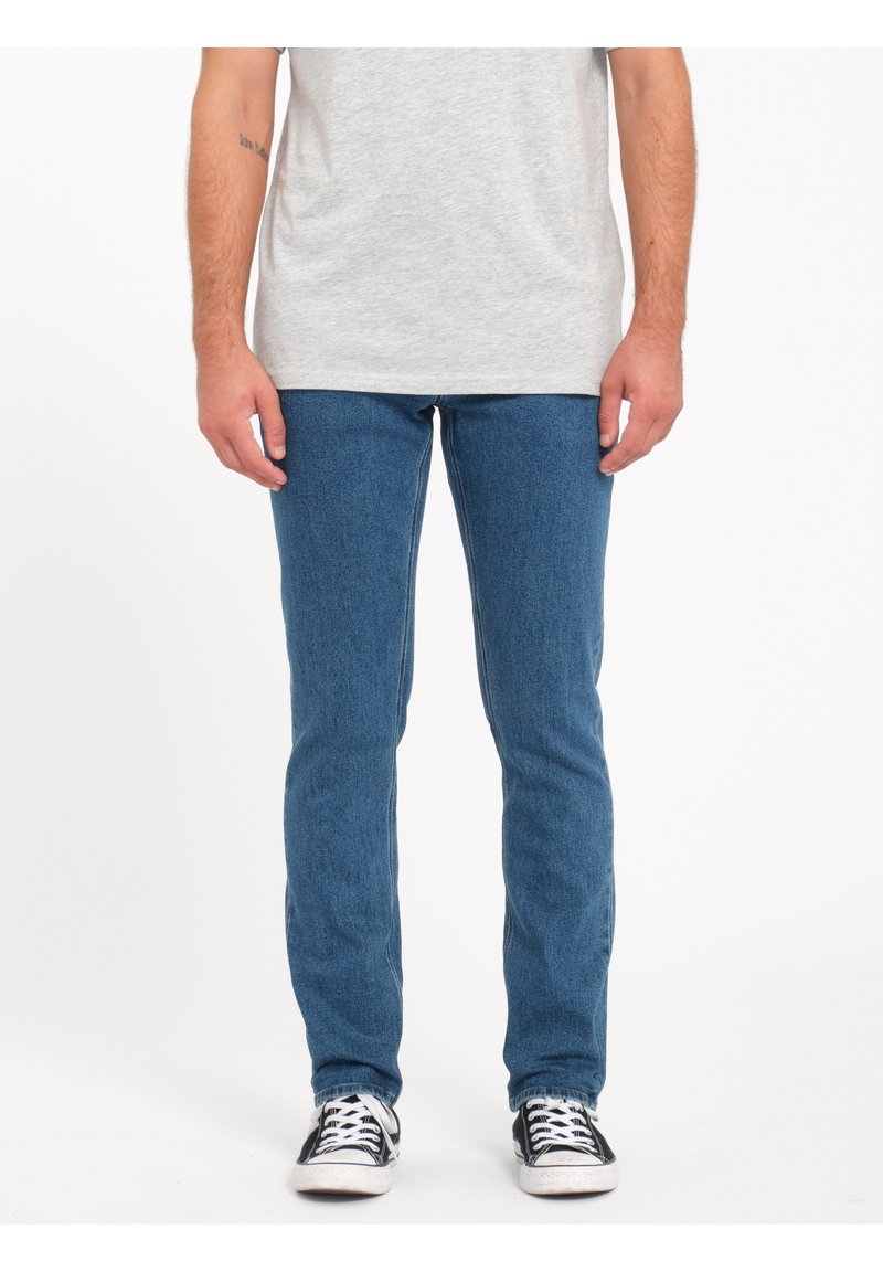 Vaqueros de mezclilla en azul con un corte slim, que presentan un diseño clásico de cinco bolsillos. Combinados con zapatillas de lona en blanco y negro.