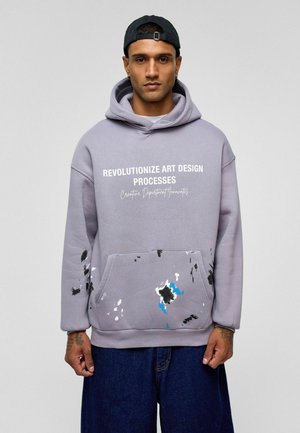 Grijze hoodie met witte grafische tekst, voorzien van een voorkzak. Accentkleuren van zwarte en blauwe verfspetters op het zakgebied. Zachte textuur.