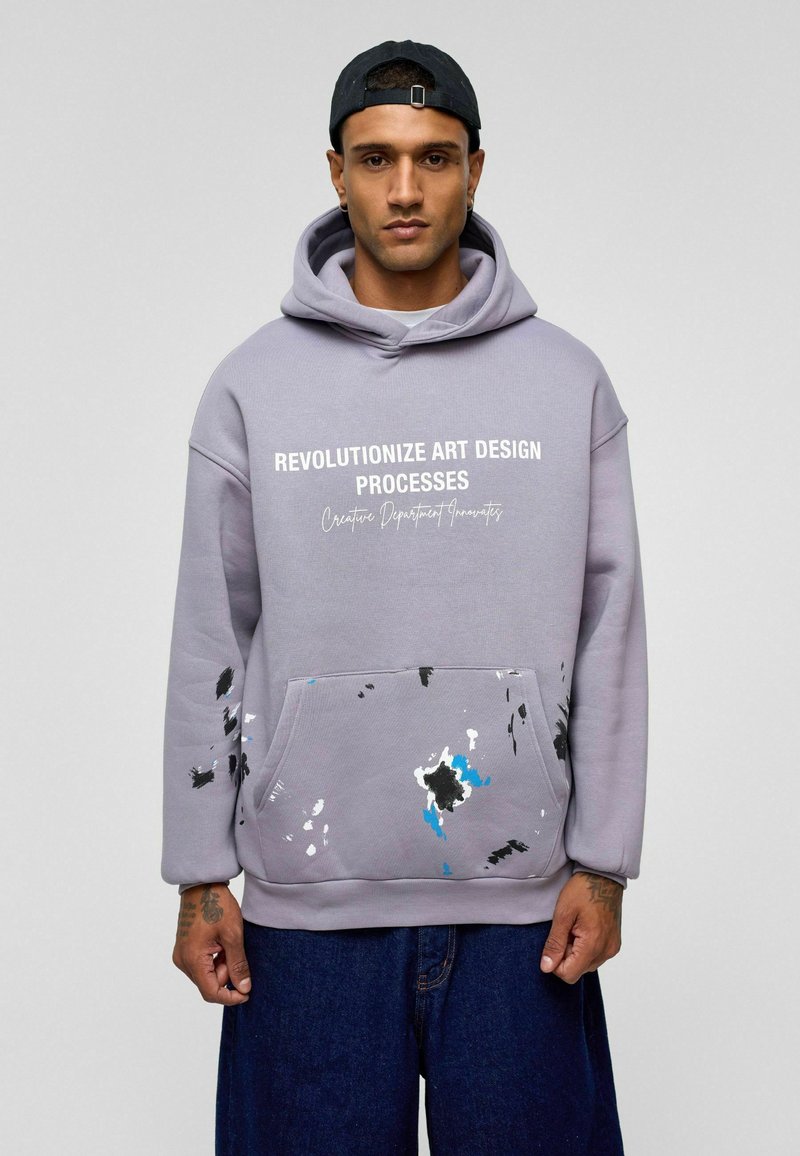 Grijze hoodie met witte grafische tekst, voorzien van een voorkzak. Accentkleuren van zwarte en blauwe verfspetters op het zakgebied. Zachte textuur.