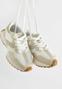 Beżowe i białe sneakersy z siateczkowym i zamszowym materiałem, okrągłym czubkiem, białymi sznurówkami i teksturowaną brązową gumową podeszwą. Akcentujące łatki po bokach.