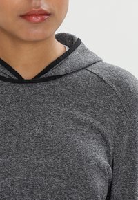 Partie inférieure du visage et haut du torse vêtus d'un sweat à capuche en maille grise avec un bord noir sur la capuche, sur un fond clair uni.