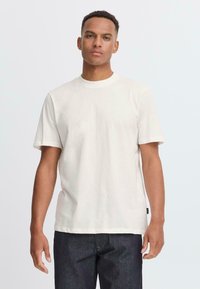 Camiseta de manga corta y cuello redondo en tela de algodón color beige claro. Diseño sencillo con corte recto y detalles mínimos. Combinada con jeans de mezclilla oscuros.