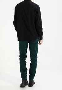 Chemise noire à manches longues avec une coupe droite, associée à un pantalon ajusté vert foncé. La chemise possède des poignets à boutons, et les deux pièces sont légères.
