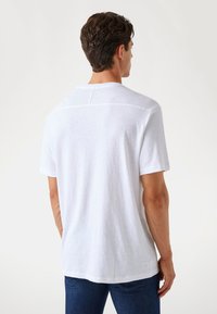 Weißes T-Shirt mit kurzen Ärmeln und Rundhalsausschnitt, weicher Textur, mit einem subtilen Nahtdetail über den Rücken, kombiniert mit blauen Jeans.