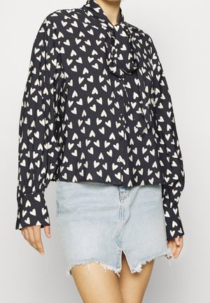 Zwarte blouse met lange mouwen en wit hartjespatroon, met een strik bij de hals, gecombineerd met een lichtblauwe, afgebladderde denim minirok met een split aan de voorkant.