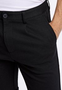 Svarta byxor med ett slätt, strukturerat tyg, smal passform, standardfickor och en enda knappstängning i midjan.