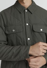 Prošívaná zelená bunda s knoflíkovým zapínáním, stříbrnými doplňky, dvěma náprsními kapsami a viditelnými diagonálními švy.