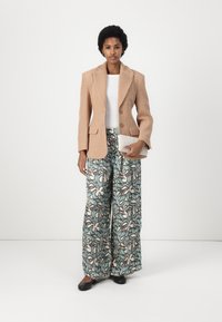 Béžový texturovaný blazer přes bílý top, spárovaný s širokými kalhotami s vzorem v modré a krémové barvě. Modelka drží psaníčko a má na sobě černé boty.