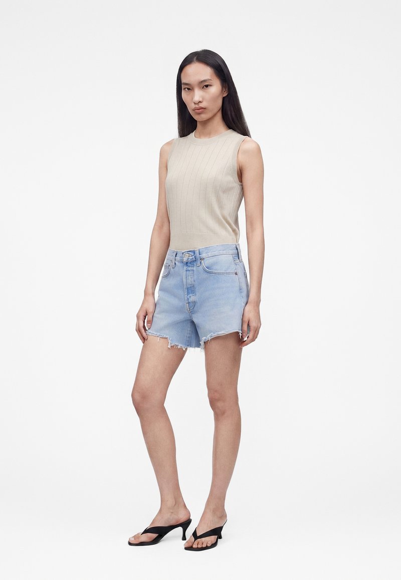 Femme portant un haut sans manches côtelé beige, un short en jean bleu clair effiloché et des sandales à talons noires, debout devant un fond blanc.