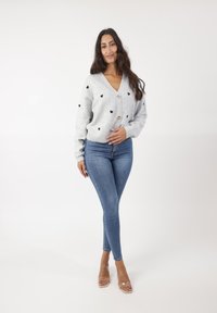 Szary, dziany kardigan z czarnymi sercami i złotymi guzikami, zestawiony z niebieskimi skinny jeansami i przezroczystymi sandałami na obcasie.