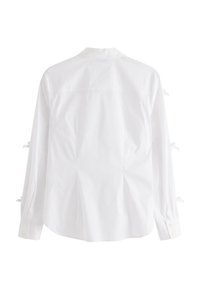Camicia bianca a maniche lunghe con colletto a punta e laccetti sulle maniche. Realizzata in cotone; presenta una spallina e dettagli arricciati.