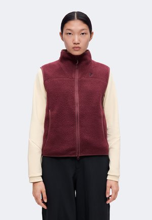 Jeune femme aux cheveux noirs porte une veste polaire bordeaux zippée par-dessus un t-shirt à manches longues crème et un pantalon noir, sur un fond uni.