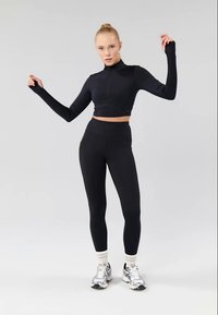 Crna uska kratka zip majica s dugim rukavima u kombinaciji s crnim uskim tajicama, koja se odlikuje modernim dizajnom i rastezljivim materijalom, u paru sa srebrnim sportskim tenisicama.
