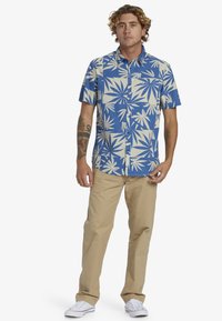Camisa de manga corta abotonada en azul con un patrón de hojas crema. Combinada con pantalones beige y zapatillas blancas. Ajuste casual con cuello.