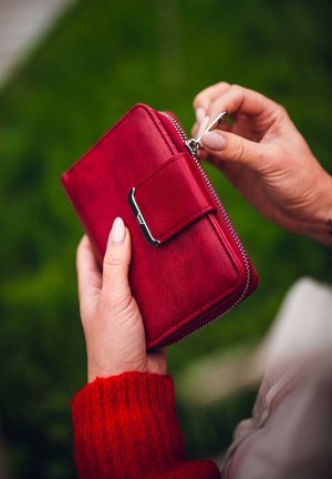 Červená kožená peněženka se zipovým uzávěrem a popruhem na druk, se stříbrným kováním, držena v rukou na zeleném pozadí.