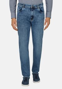 Mann trägt mittelblaue Jeans mit geradem Bein, graues Langarmshirt und dunkelblaue Sneaker mit weißen Sohlen, steht mit entspannten Händen.