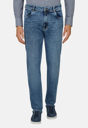 Mann trägt mittelblaue Jeans mit geradem Bein, graues Langarmshirt und dunkelblaue Sneaker mit weißen Sohlen, steht mit entspannten Händen.