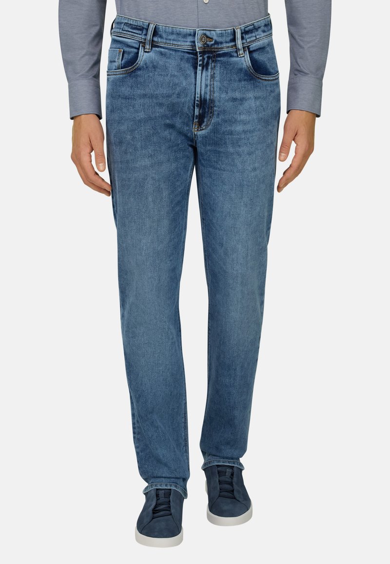 Mann trägt mittelblaue Jeans mit geradem Bein, graues Langarmshirt und dunkelblaue Sneaker mit weißen Sohlen, steht mit entspannten Händen.