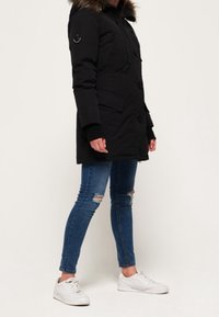 Svart parkacoat med foder av päls i huvan, med flera knappar, patchdetaljer och stora fickor. Kombinerad med tajta jeans och vita sneakers.